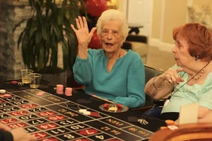Two elderly women enjoy Casino Night at a roulette table; one raises her hand and smiles while the other points, with chips, drinks, and a plate of snacks on the table.
