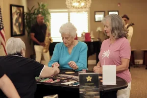 Three older women play cards at a Casino Night table in a well-lit room, while two musicians perform in the background.