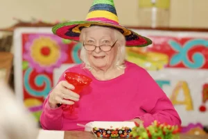 An elderly woman wearing a colorful sombrero and glasses holds a red drink, celebrating Cinco de Mayo at a table decorated with bright festive patterns.