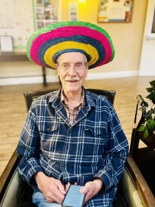 An elderly man wearing glasses and a colorful sombrero sits in a chair, holding a smartphone, dressed in a checkered shirt, as he enjoys the festive spirit of Cinco de Mayo with a blurred background behind him.
