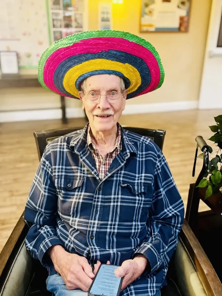 An elderly man wearing glasses and a colorful sombrero sits in a chair, holding a smartphone, dressed in a checkered shirt, as he enjoys the festive spirit of Cinco de Mayo with a blurred background behind him.