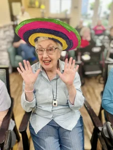 An elderly woman wearing a colorful sombrero sits and smiles with her hands raised, celebrating Cinco de Mayo, surrounded by other people in a lively community room.