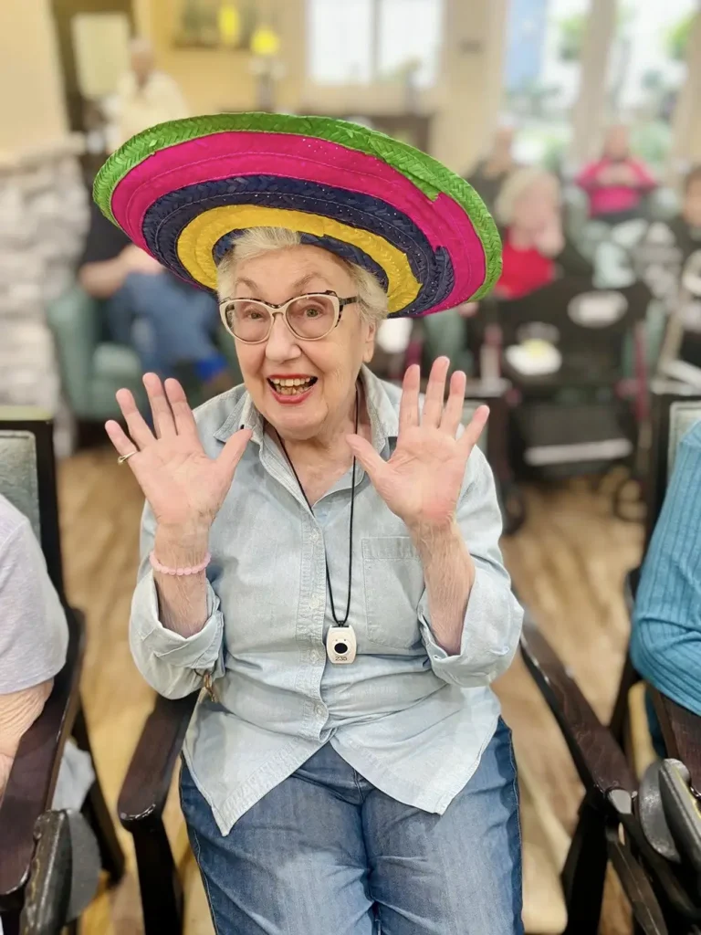 An elderly woman wearing a colorful sombrero sits and smiles with her hands raised, celebrating Cinco de Mayo, surrounded by other people in a lively community room.