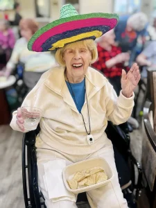 Elderly woman in a wheelchair wearing a colorful sombrero, smiling, holding a drink and a tray of chips, celebrating Cinco de Mayo with people and a festive atmosphere in the background.