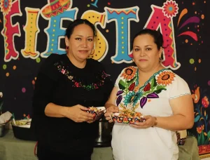Two women stand in front of a colorful "FIESTA" sign, each holding a plate of food. Dressed in traditional embroidered blouses, they celebrate Cinco de Mayo with vibrant smiles and festive spirit.