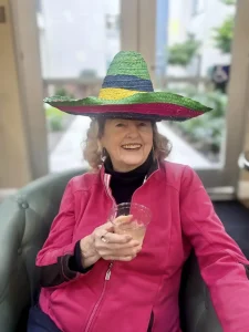 Older woman in a bright pink jacket sits on a green chair, smiling and holding a drink, wearing a large, colorful sombrero—celebrating Cinco de Mayo in festive style.