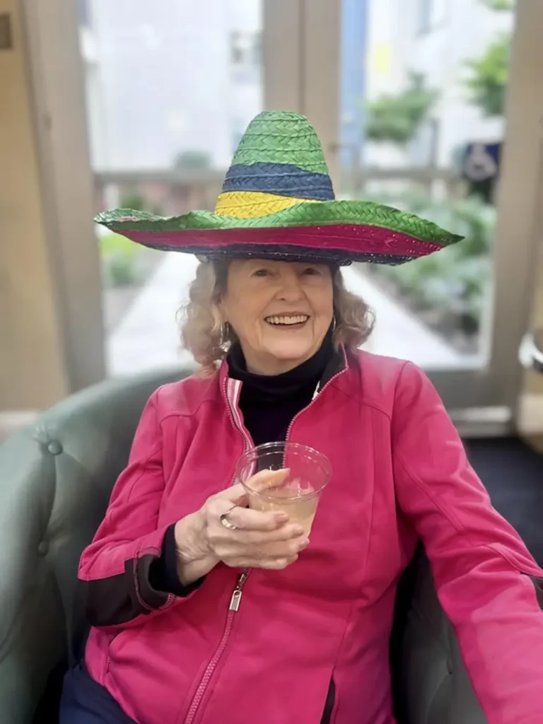 Older woman in a bright pink jacket sits on a green chair, smiling and holding a drink, wearing a large, colorful sombrero&mdash;celebrating Cinco de Mayo in festive style.
