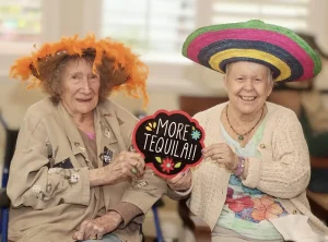 Two elderly women wearing festive hats sit together indoors, smiling and holding a sign that says "MORE TEQUILA!!" as they celebrate Cinco de Mayo.