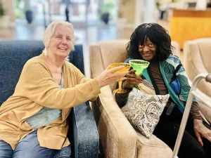 Two older women sit on a couch, smiling and holding up colorful drinks in a toast as they celebrate Cinco de Mayo.