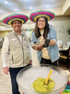 Two people wearing colorful sombreros stand together—one smiling while holding a bowl of chocolate—as they celebrate Cinco de Mayo. A large bowl of guacamole with a spoon sits in the foreground, adding to the festive Mexican holiday vibe.