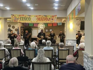 A mariachi band performs lively tunes on stage for an audience of elderly people celebrating Cinco de Mayo in a festively decorated community room.