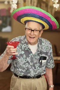 An older man wearing glasses and a colorful sombrero celebrates Cinco de Mayo, holding up a red drink in a festive setting, dressed in a floral shirt and khaki pants.