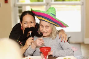 Two women pose together at a table on Cinco de Mayo; one wears a colorful sombrero, and the other holds a fake mustache on a stick. Food and a drink are visible, adding to the festive May 5th celebration.