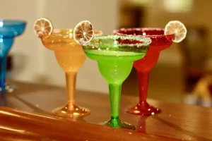 Three colorful margarita glasses—orange, green, and red—each garnished with a lime slice and a salted rim, are displayed on a wooden surface, perfect for celebrating Cinco de Mayo.