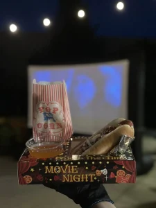 A tray with a hot dog, popcorn, and dipping sauces is held up in front of an open-air movie screen at night. "Movie Night" is printed on the tray, capturing the magic of an outdoor cinema experience.