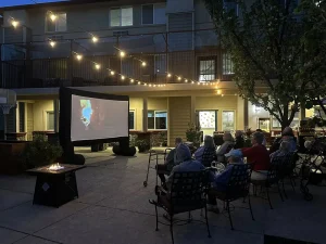 A group of people sit in chairs outdoors at night watching a Cinema Under the Sky movie projected on a screen, with string lights overhead and a fire pit nearby.