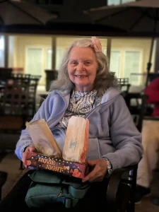 An elderly woman with gray hair and a flower hair clip smiles while holding an empty "Movie Night" popcorn box in her lap. Seated indoors in a light purple jacket, she looks ready for another Cinema Under the Sky experience.