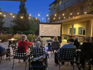 A group of seniors sit outdoors in chairs and wheelchairs, enjoying a Cinema Under the Sky as they watch a movie on a large screen at night beneath twinkling string lights.