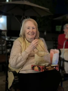 Elderly woman sitting outdoors, smiling as she enjoys Cinema Under the Sky, holding a popcorn bag and a "Movie Night" box on her lap.