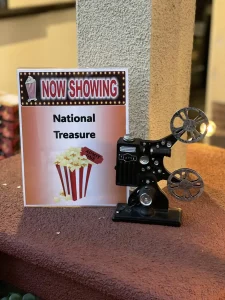 A sign reads "Now Showing: National Treasure" beside a small vintage film projector, both placed on a red carpeted step—perfect for a magical Cinema Under the Sky experience.