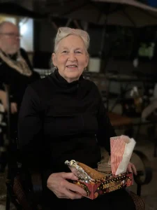 An older woman in a black top sits holding a tray labeled "Movie Night," filled with popcorn and snacks, smiling warmly. Glasses rest on her head as she enjoys the Cinema Under the Sky; another person is blurred in the background.