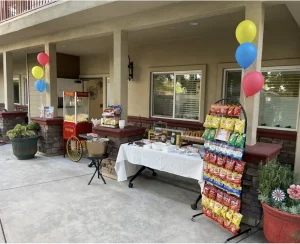 Outdoor snack stand setup for Cinema Under the Sky with a popcorn machine, tables of food, a display rack of chips, and colorful balloons arranged in front of a building.