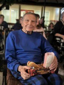 An elderly man in a blue sweatshirt and plaid pants sits outside at Cinema Under the Sky, holding a tray with a hot dog and popcorn labeled "Movie Night," as people are seated in the background.