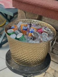 A metal bucket filled with ice and assorted canned sodas—Fanta, Sprite, A&W Root Beer, and Canada Dry—sits on a round black table outdoors, ready for a refreshing Cinema Under the Sky experience.