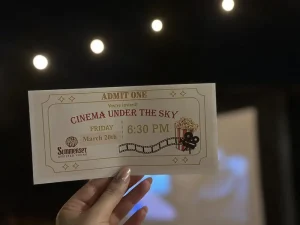A hand holds a "Cinema Under the Sky" ticket for Friday, March 20th at 6:30 PM, set against blurred lights and a glowing screen in the background.