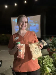 A smiling woman in orange scrubs holds a can of soda, popcorn, and a soft pretzel, enjoying Cinema Under the Sky as a movie is projected on an outdoor screen at night.