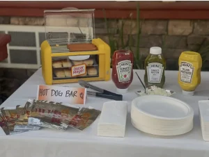 A hot dog bar setup for Cinema Under the Sky features buns in a warmer, ketchup, relish, mustard, wrapped hot dogs, paper plates, napkins, and utensils on a white tablecloth.