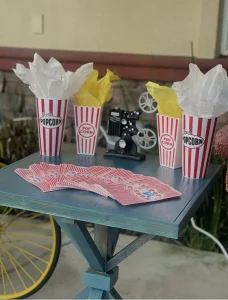 A small blue table holds striped popcorn containers with tissue paper, a miniature film projector, and a spread of red playing cards—perfect touches for an open-air movie or cozy Cinema Under the Sky gathering.