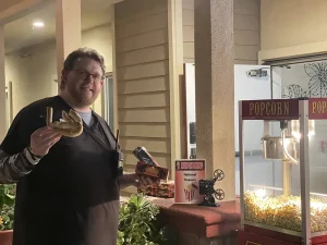 A man stands outside next to a popcorn machine, holding a gold painted object and a box of snacks, with a "National Treasure" movie sign on the table, ready for Cinema Under the Sky.