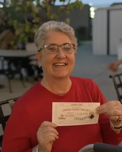 A person with short gray hair and glasses, wearing a red shirt, smiles while holding up an "open air movie" Cinema Under the Sky event ticket outdoors.