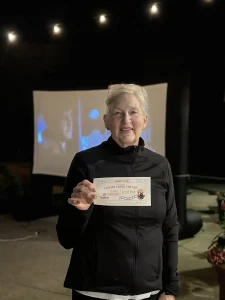 An older woman wearing glasses and a black jacket stands outdoors at night, holding a ticket at Cinema Under the Sky. In the background, a large screen displays an image as part of this open-air movie experience.