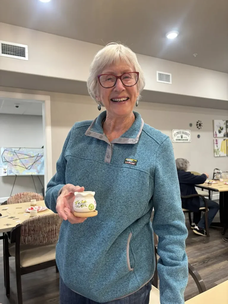 An older woman wearing glasses and a blue fleece stands indoors, smiling and holding a small jar with a nature-themed label. Other people are seated at tables in the background.