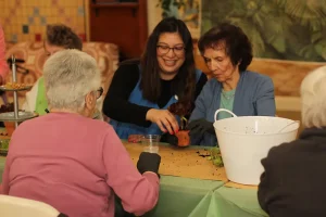 A group of women sit around a table, working together to pot plants. Soil and gardening supplies are spread on the table.