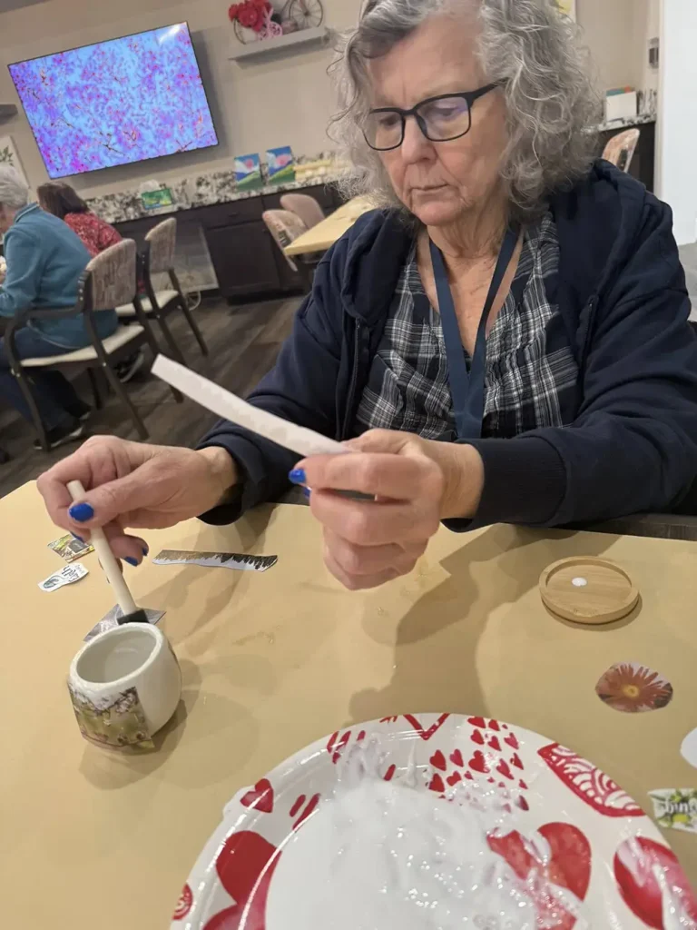 An older woman sits at a table holding a craft stick and paper, with craft supplies and an empty plate in front of her.