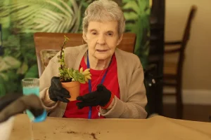 An elderly woman wearing black gloves holds a small potted succulent while sitting at a table with soil scattered on the surface.