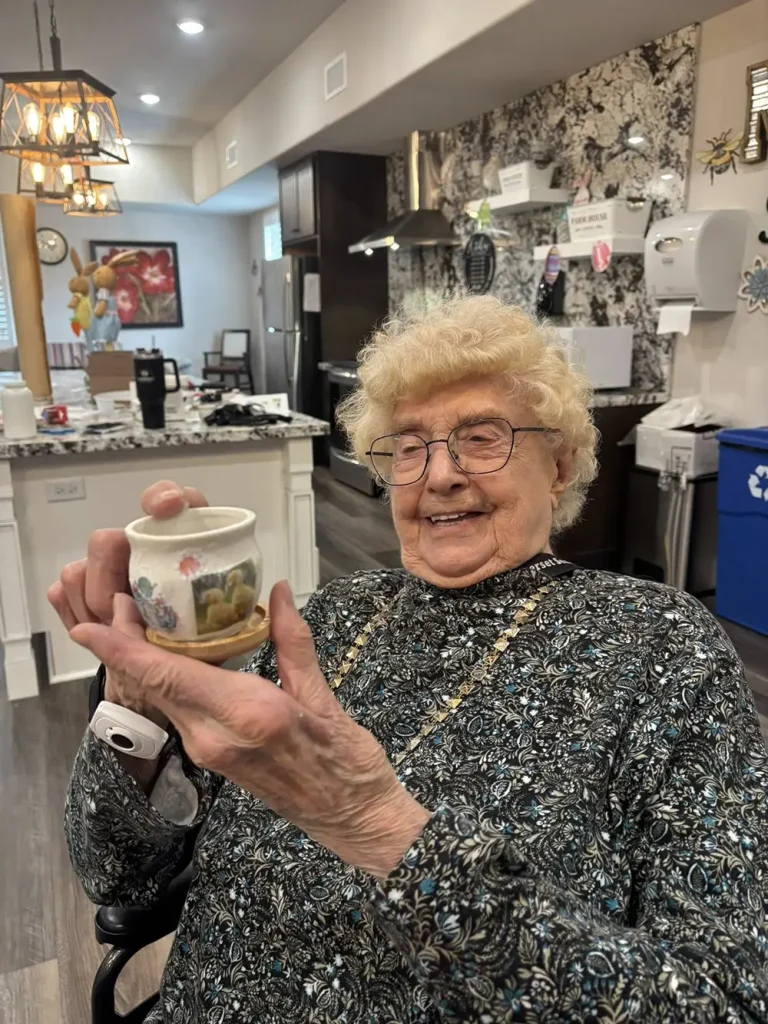 An elderly woman with glasses and curly hair smiles while holding a small painted cup in a modern kitchen.