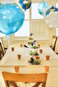 A dining table set with small flower pots, a tiered plant arrangement, and blue balloons, in a bright room with wooden chairs and large windows.