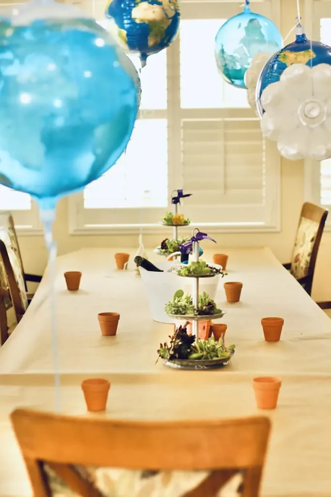 A dining table set with small flower pots, a tiered plant arrangement, and blue balloons, in a bright room with wooden chairs and large windows.