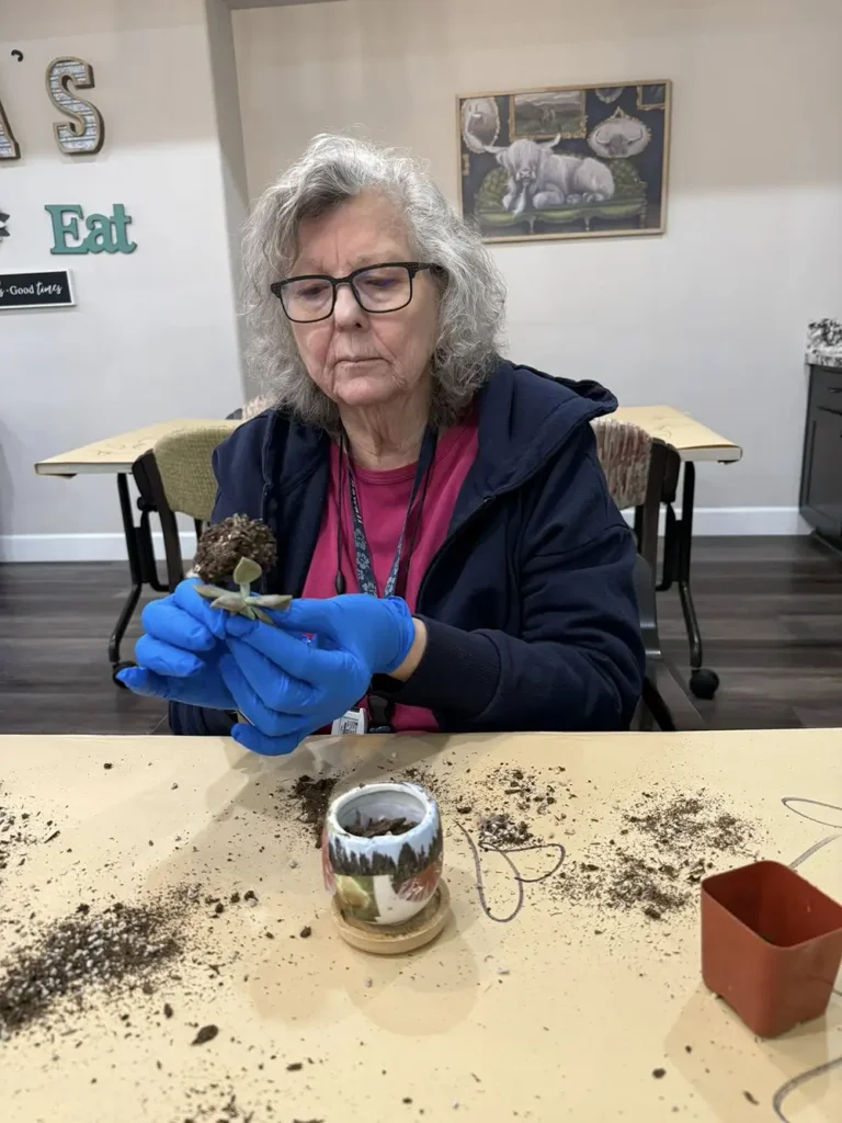 An older woman wearing blue gloves repots a small plant at a table, with soil and gardening tools scattered around. A mug and small pot are also on the table.