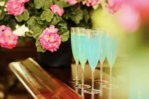 Six champagne flutes filled with blue liquid are arranged on a polished wooden surface beside a pot of pink flowers.