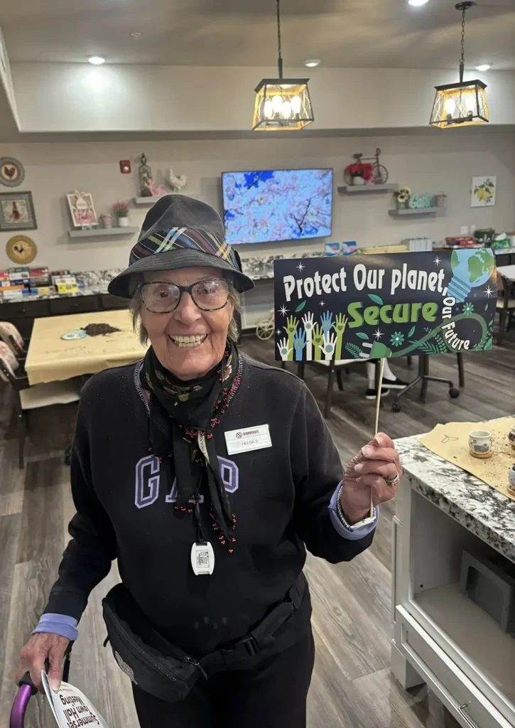 An elderly woman in a hat and glasses holds a sign reading "Protect Our planet, Secure our future" in a decorated room.