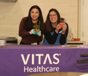 Two women stand behind a table with a VITAS Healthcare cloth, each holding a potted plant and smiling at the camera.