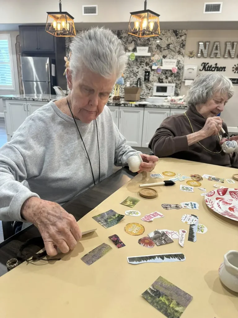 Two elderly women sit at a table working on a crafts project with stickers and paper in a well-lit kitchen area.