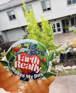A hand holds a colorful sign reading "The rotation of the Earth really makes my day" in front of a courtyard with greenery and a building in the background.