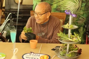 An older man wearing glasses and gloves sits at a table, smiling while potting a succulent plant. A tiered tray with more succulents and craft items are on the table.