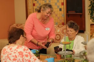 A woman assists two elderly women with planting succulents at a table in a communal indoor setting.